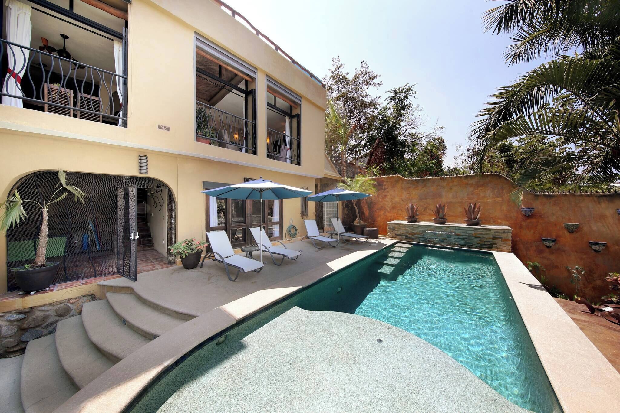 Une piscine avec des chaises longues et des parasols, un immeuble à plusieurs étages doté de balcons et un mur en pierre.