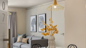 Ein modernes Wohnzimmer mit einem grauen Sofa, einem Glastisch mit schwarzen Stühlen, einer gelben Pendelleuchte und gerahmten Kunstwerken an der Wand.