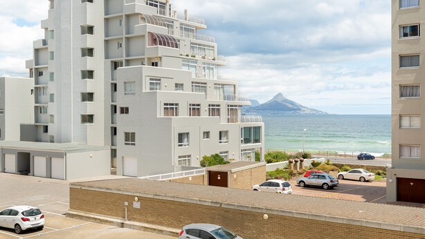 Un edificio residencial de varios pisos con balcones, un estacionamiento y vista al océano y una montaña a lo lejos.