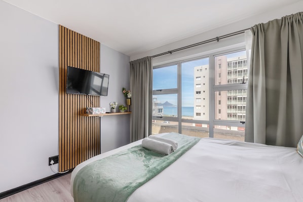 A hotel room with a bed, a TV mounted on a wooden panel, a window with curtains, and a view of buildings and the sea.