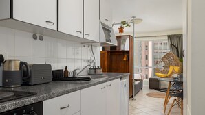 Una cocina moderna con armarios blancos, encimera de granito y electrodomésticos de acero inoxidable. Hay una zona de comedor con mesa y sillas, y una ventana con vistas a edificios.