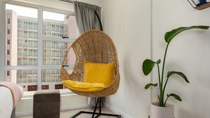 Una habitación moderna con una silla de mimbre, una planta en maceta y vistas de edificios a través de una ventana.