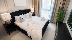 Een net opgemaakt bed met een witte deken en kussens, een bedlampje en een nachtkastje met een lamp.