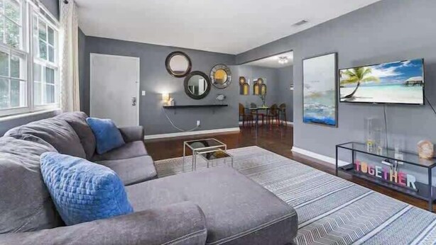 A modern living room with a grey sofa, a coffee table, a TV, and wall art. - Eclectic Escape #4 - Team Joseph Ellen (Jacksonville)