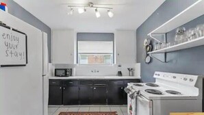 A modern kitchen with a white stove, black cabinets, a microwave, and a window with blinds. - Eclectic Escape #4 - Team Joseph Ellen (Jacksonville)