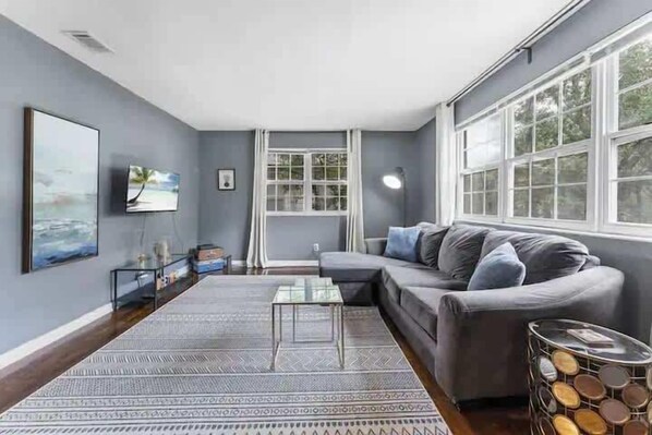 A modern living room with a grey sofa, a glass coffee table, a patterned rug, and a large window with curtains.