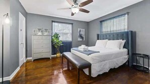 A bedroom with a bed, a ceiling fan, a dresser, a bench, a plant, and a window. - Eclectic Escape #4 - Team Joseph Ellen (Jacksonville)