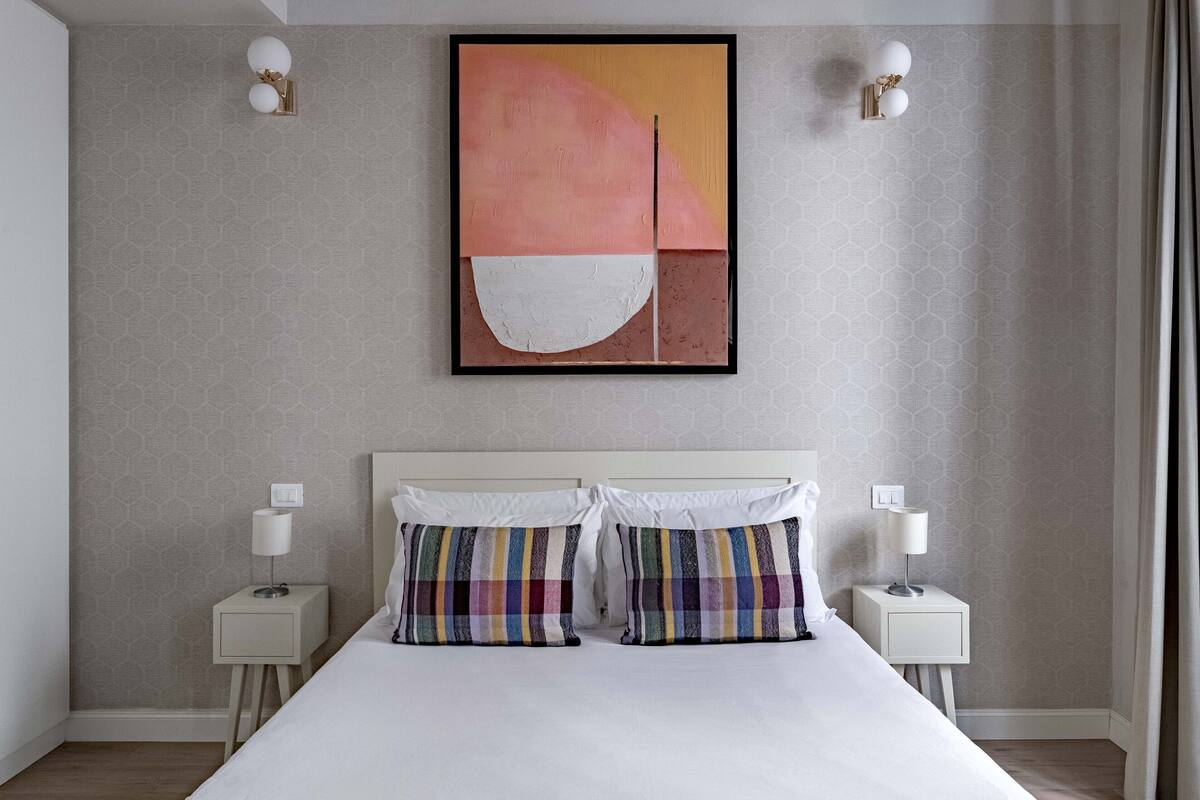 A modern hotel room with a large bed, two bedside tables with lamps, a large abstract painting on the wall, and a patterned wallpaper.