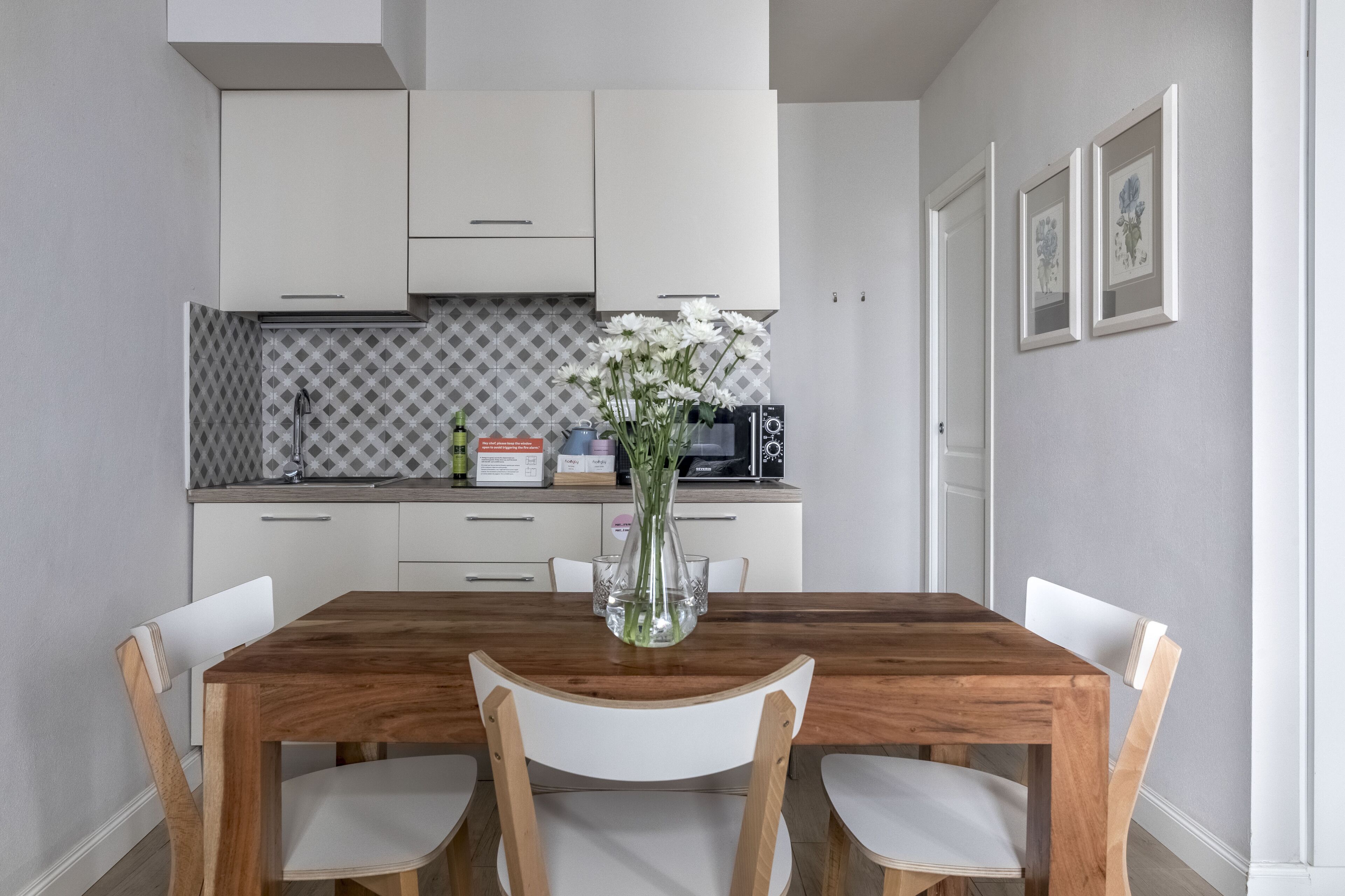 Een eethoek met een houten tafel en witte stoelen, een vaas met bloemen en een keuken op de achtergrond.