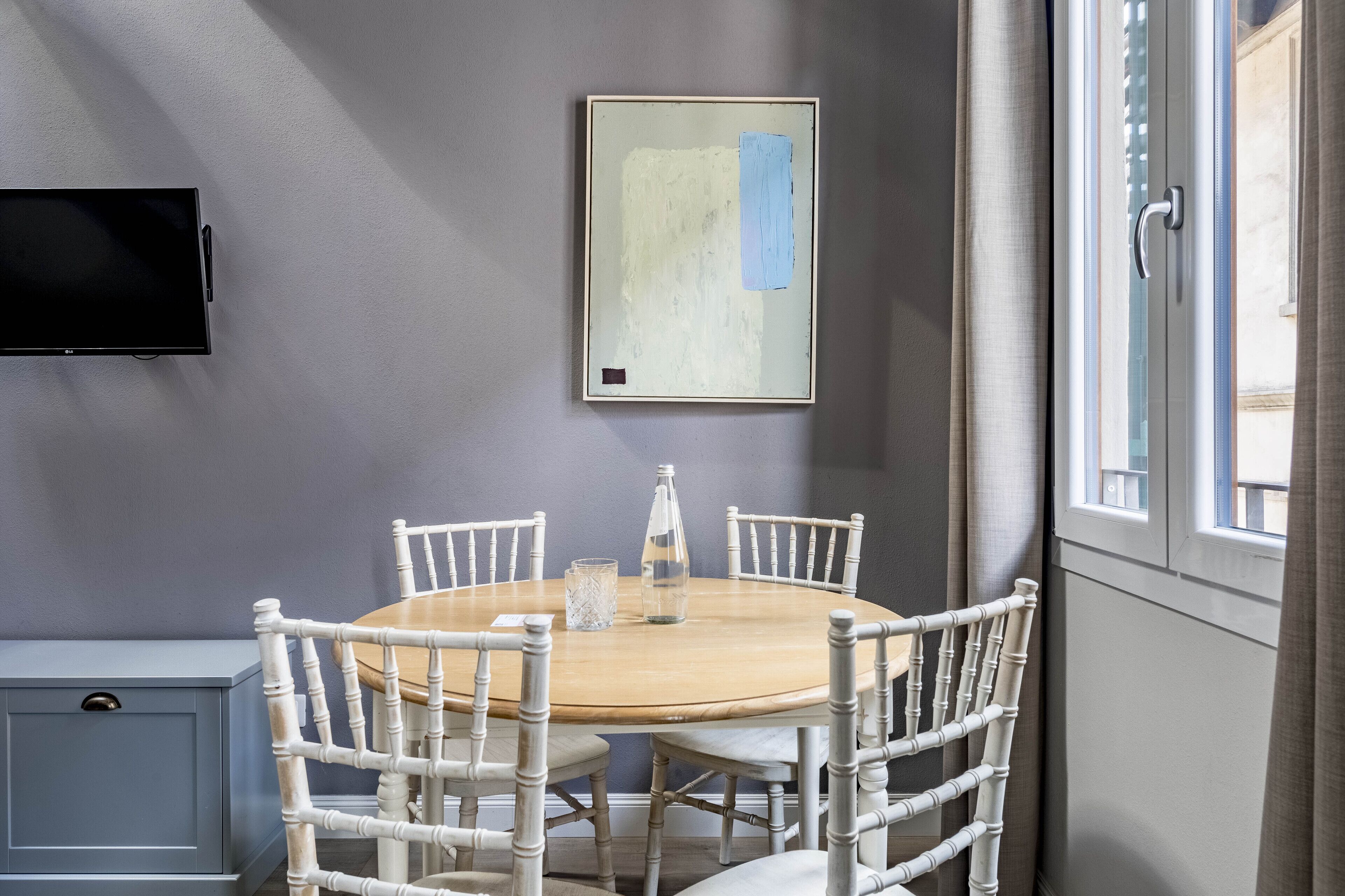 A dining area with a round table, white chairs, a TV mounted on the wall, and a window with curtains.