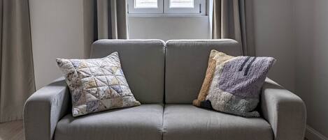 A grey sofa with two patterned cushions, a window with curtains, and a plain wall.