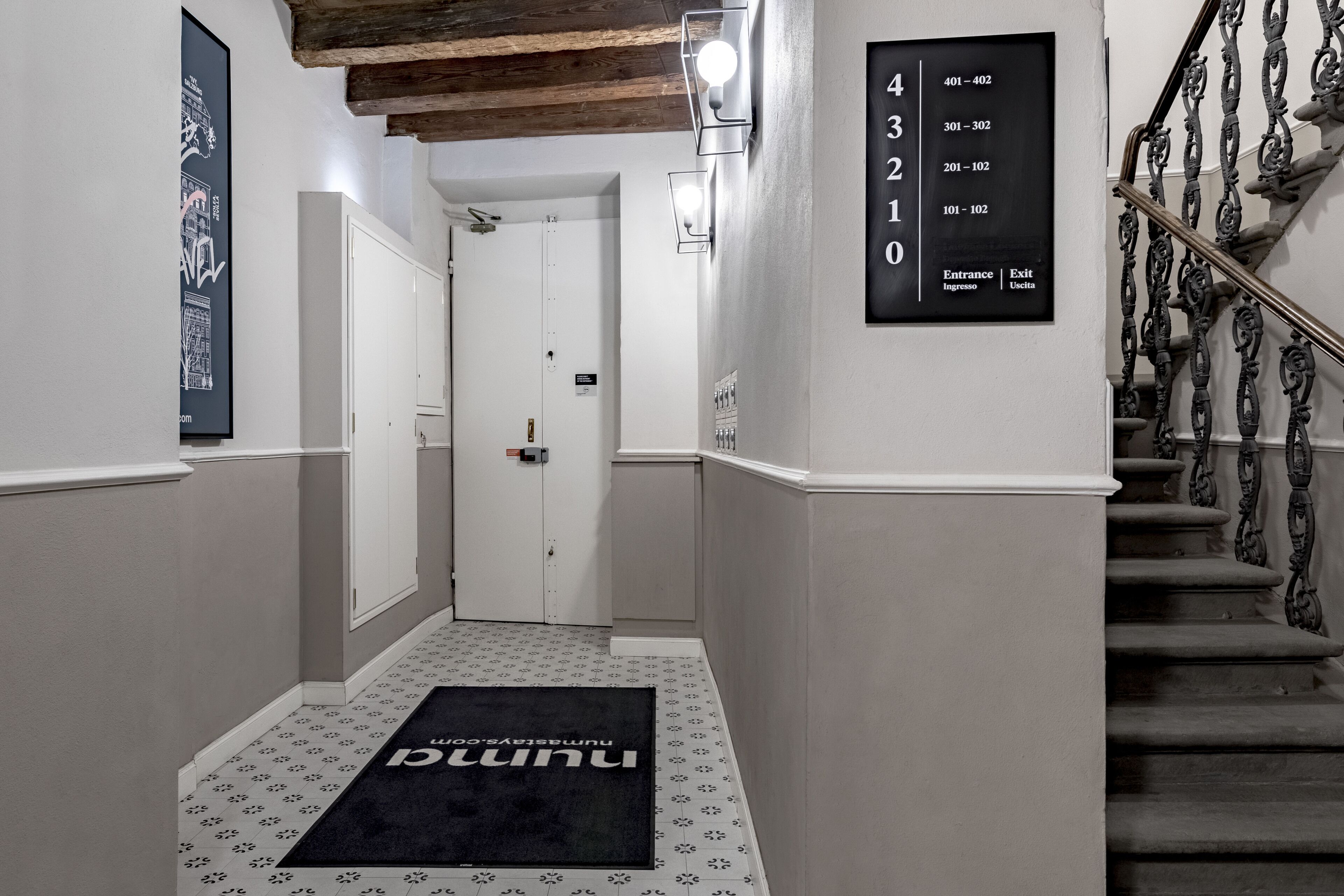 A hotel hallway with a black doormat, a staircase with ornate metal railings, and a wall-mounted directory listing room numbers and directions.