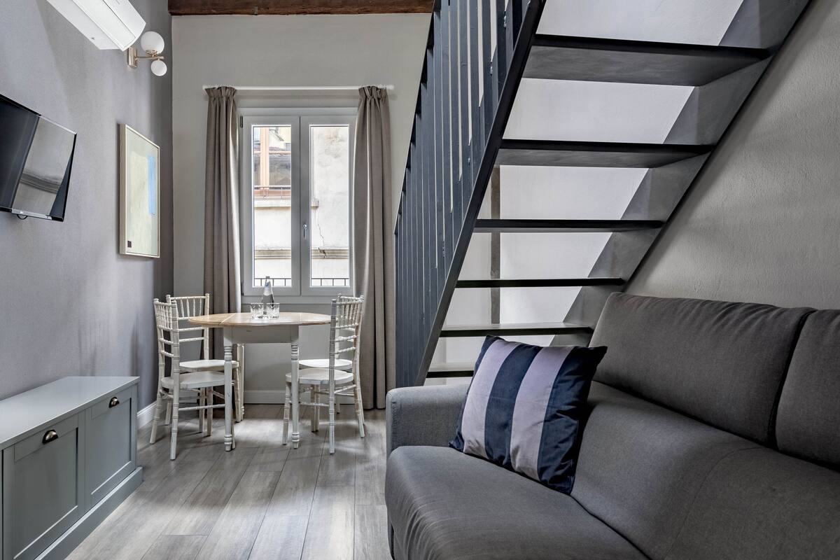 A modern loft bedroom with a sloped ceiling, a built-in desk, a dining area, and a sofa.