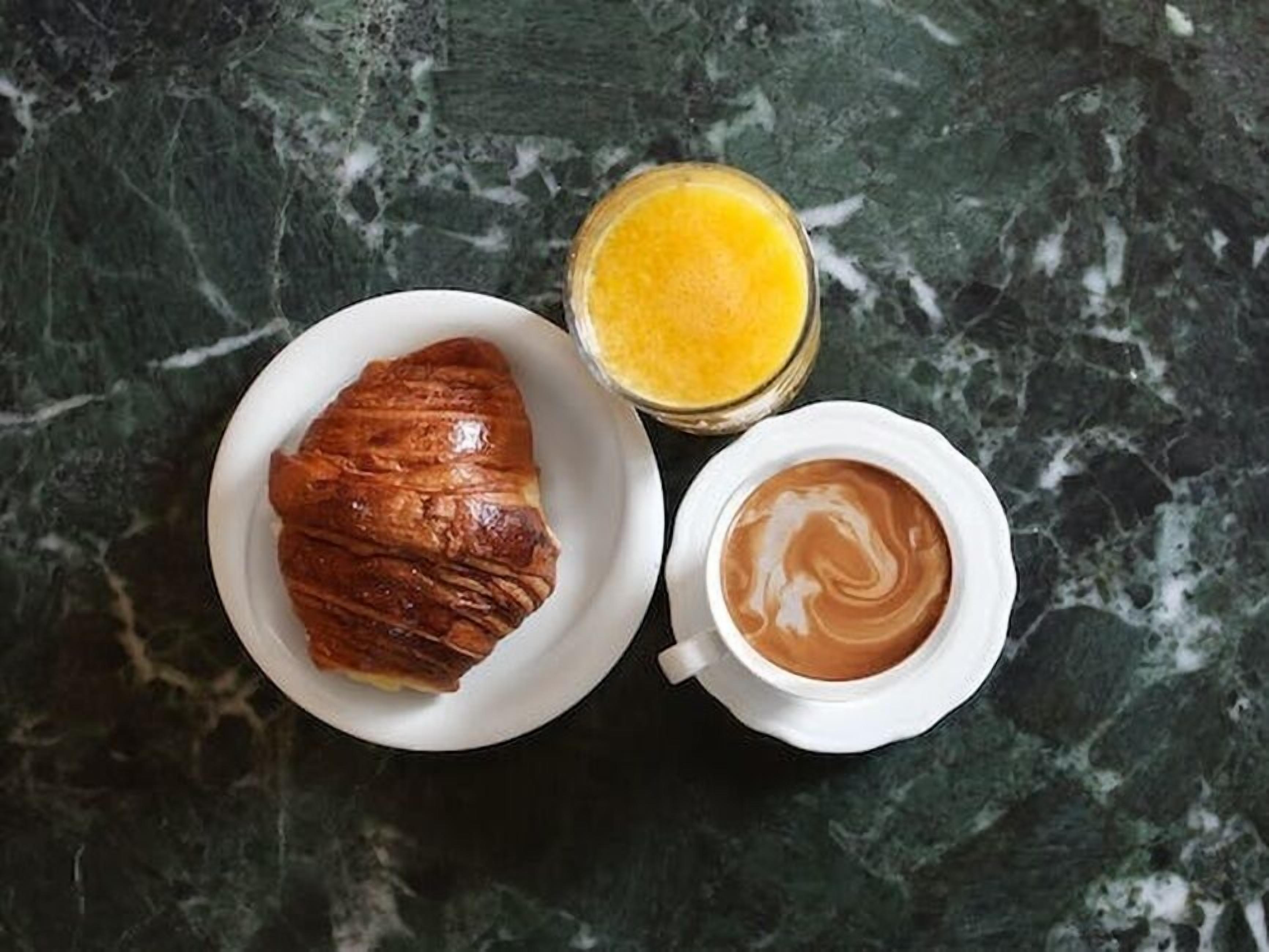 Un croissant en un plato, un vaso de zumo de naranja y una taza de café sobre una superficie de mármol.
