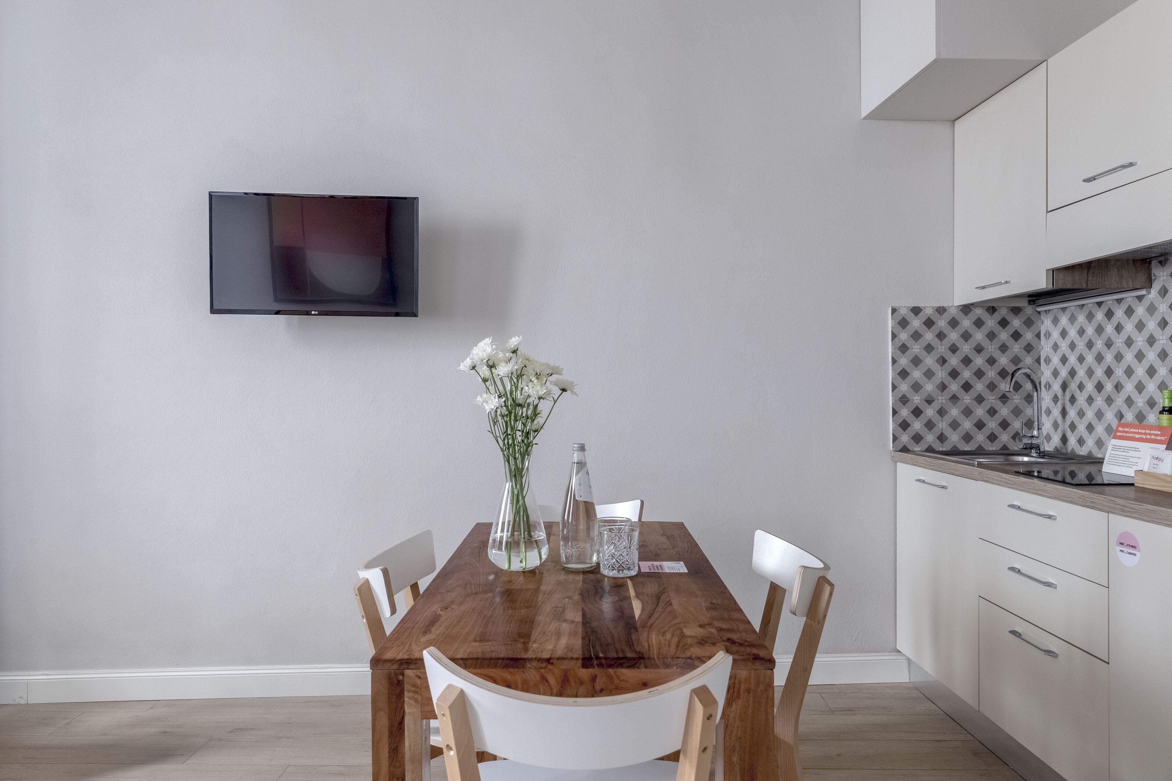 Uma cozinha moderna com uma mesa de jantar de madeira, cadeiras brancas, uma TV montada na parede e um revestimento de parede estampado.