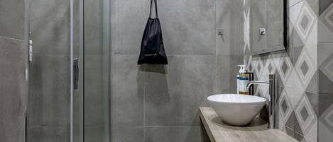 A modern bathroom with a glass shower enclosure, a wooden vanity with a white sink, and a black bag hanging on the wall.