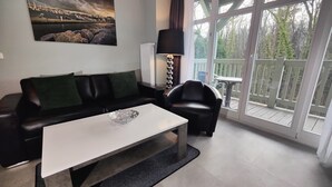 Un salon moderne avec un canapé en cuir noir, une table basse blanche et un fauteuil noir. Une grande fenêtre offre une vue sur un balcon et des arbres.