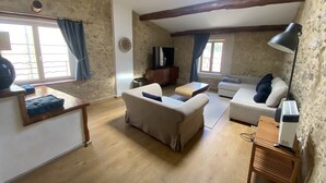 Ein rustikales Wohnzimmer mit sichtbaren Holzbalken, einer Steinwand und einem Oberlicht. Es gibt zwei Sofas, einen kleinen Tisch und einen Fernseher auf einer Holzkommode.
