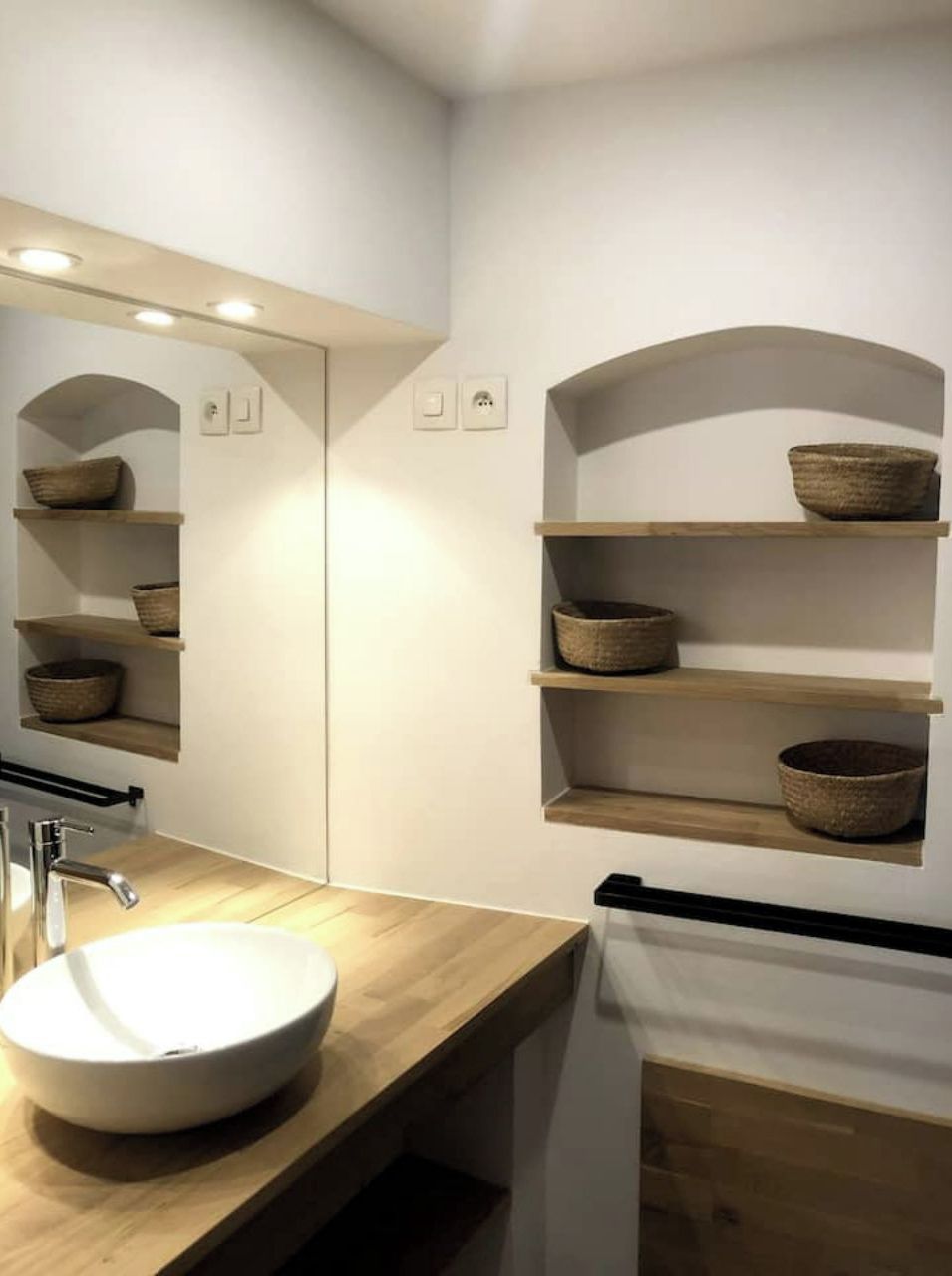 A modern bathroom with a wooden vanity, a round sink, and built-in shelves with woven baskets.