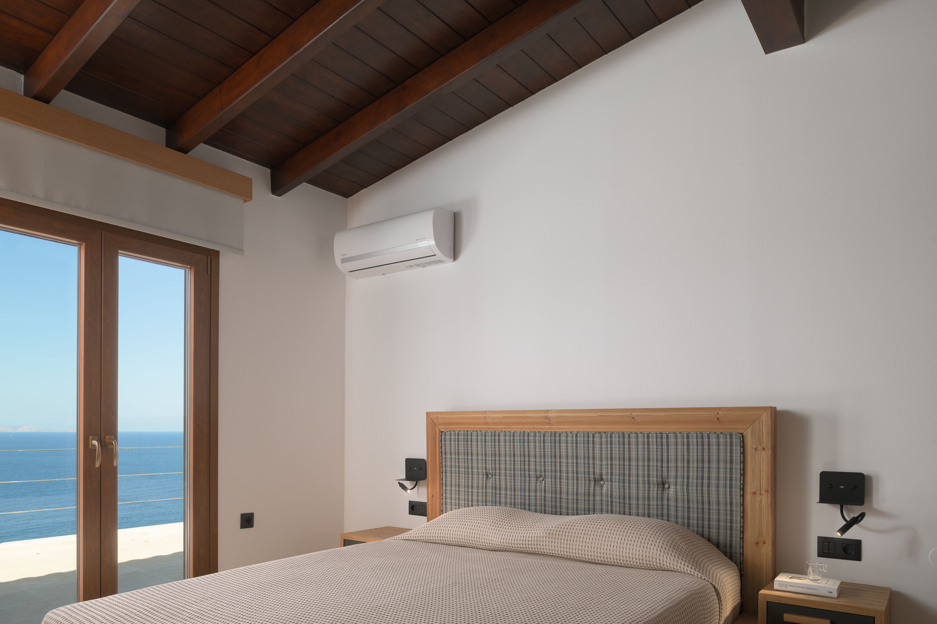 A bedroom with a wooden ceiling, a large window with a view of the sea, a bed with a checkered headboard, and a bedside table with a lamp.