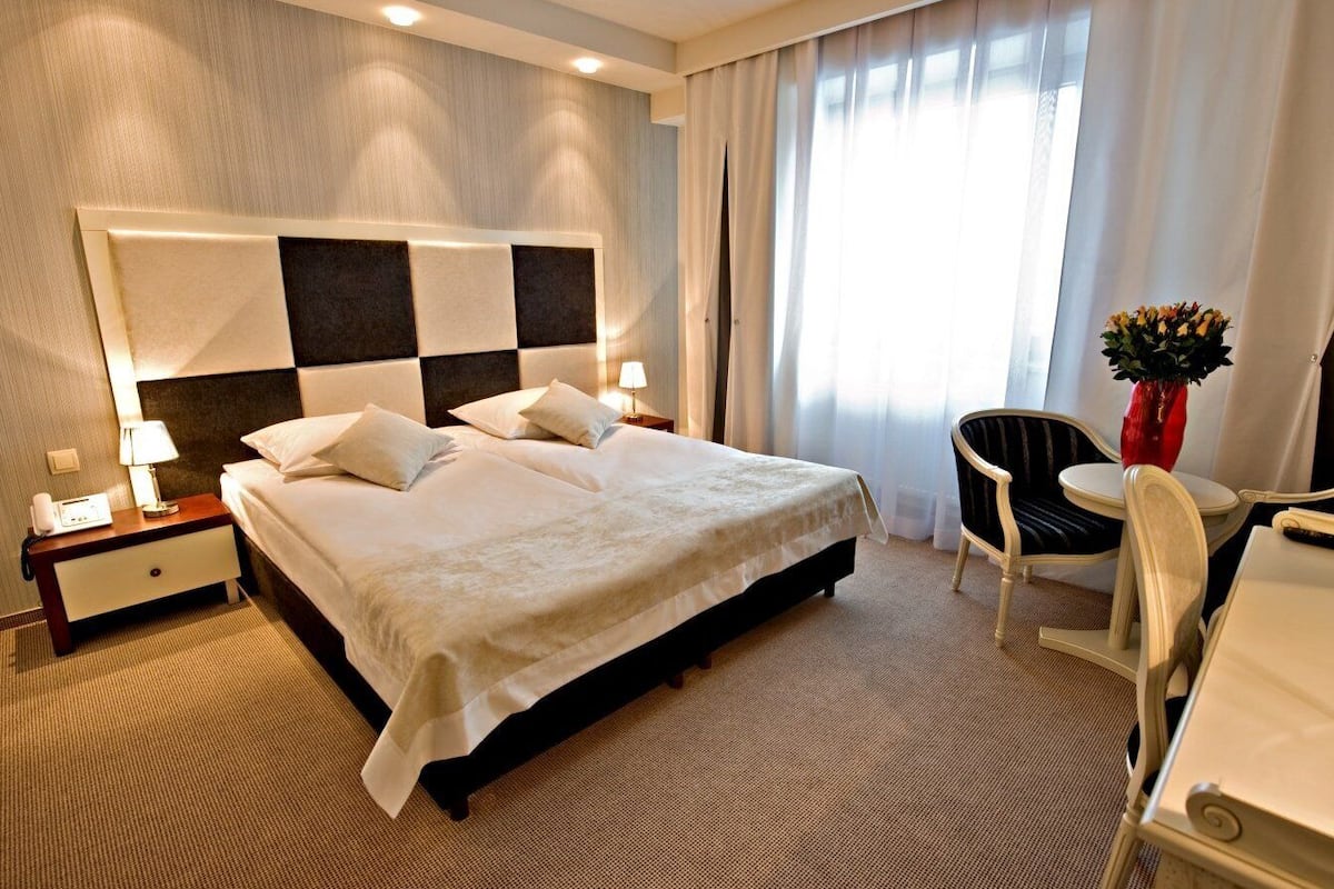 A hotel room with a large bed, a desk, a chair, and a nightstand with a lamp.
