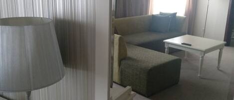 Habitación de hotel con sofá, una mesita, lámpara y vistas al exterior a través de cortinas.