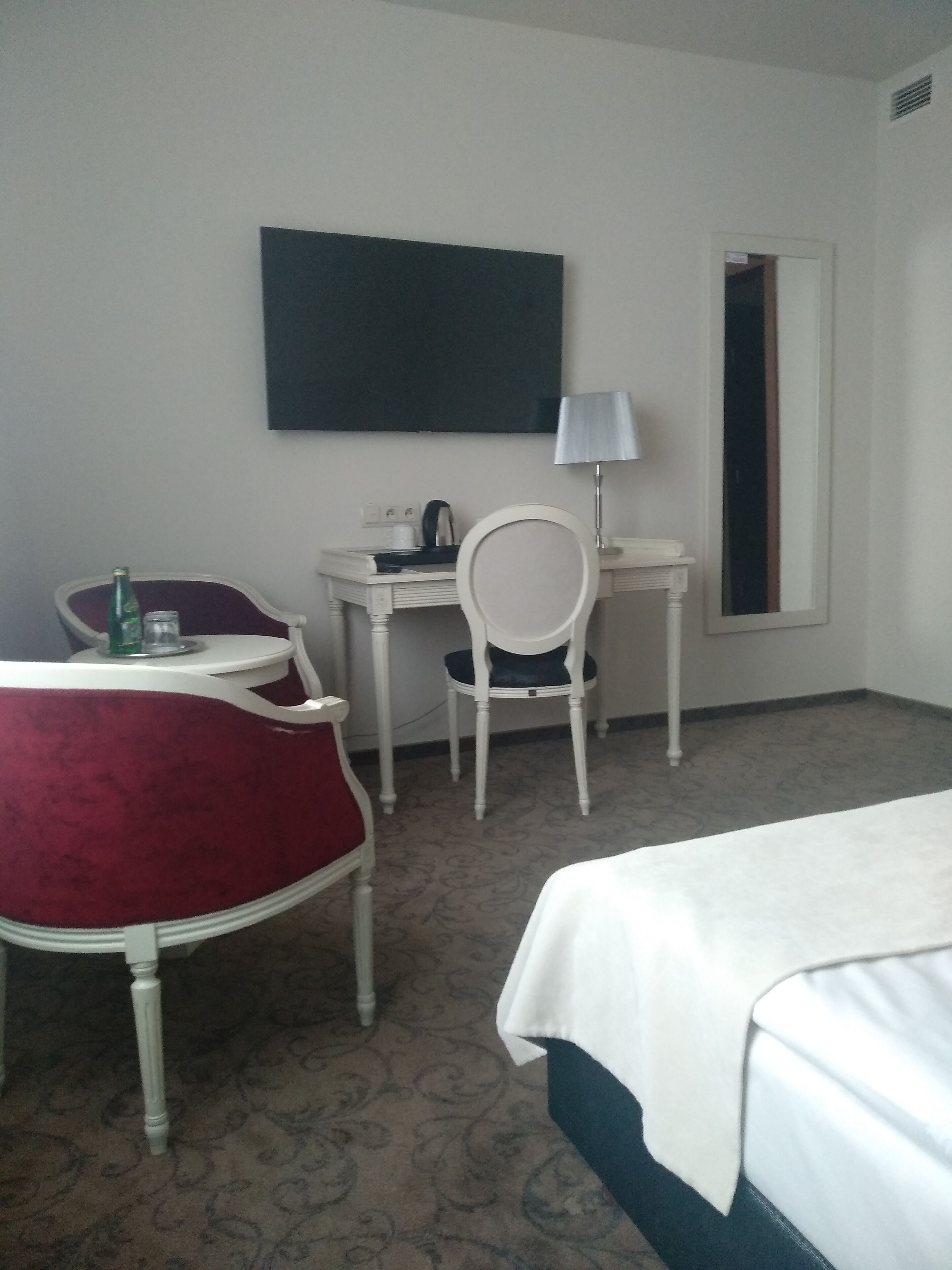A hotel room with a bed, a desk, a chair, a lamp, and a television on the wall.