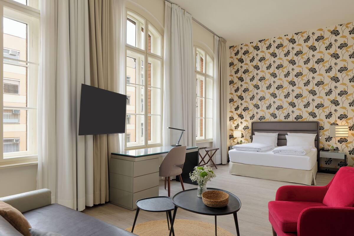 A hotel room with a bed, a TV, a sofa, a red armchair, and a small table with a vase of flowers.