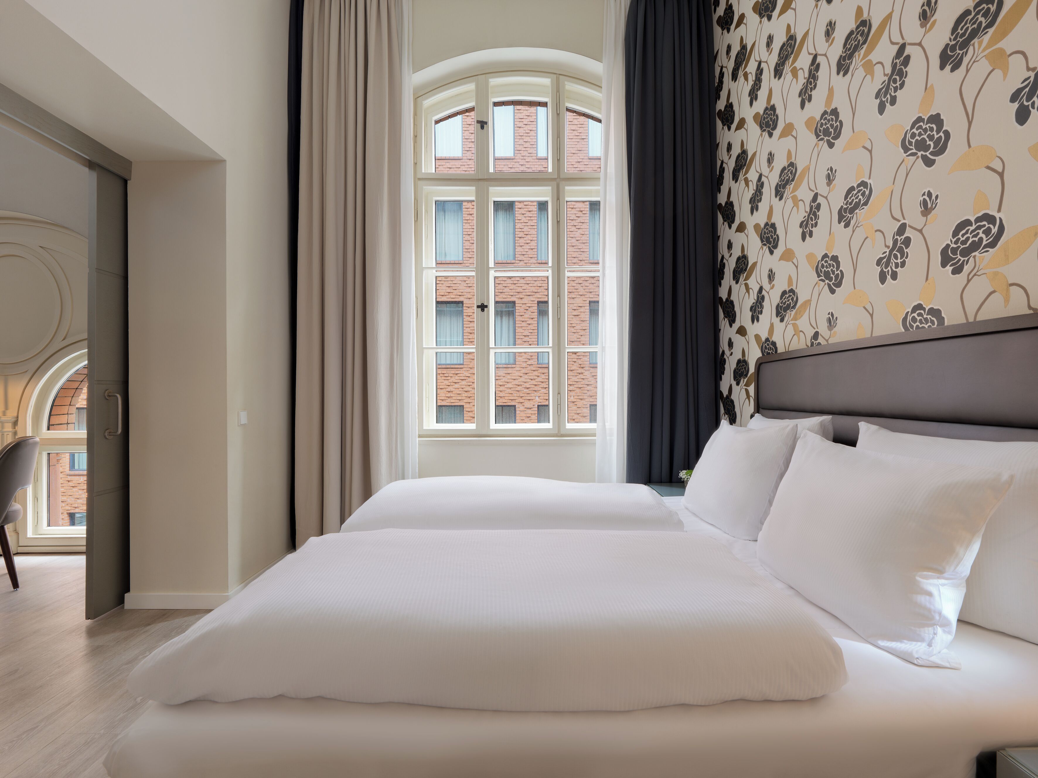Ein Hotelzimmer mit einem großen Bett, Blick auf ein Backsteingebäude und einer Wand mit Muster.