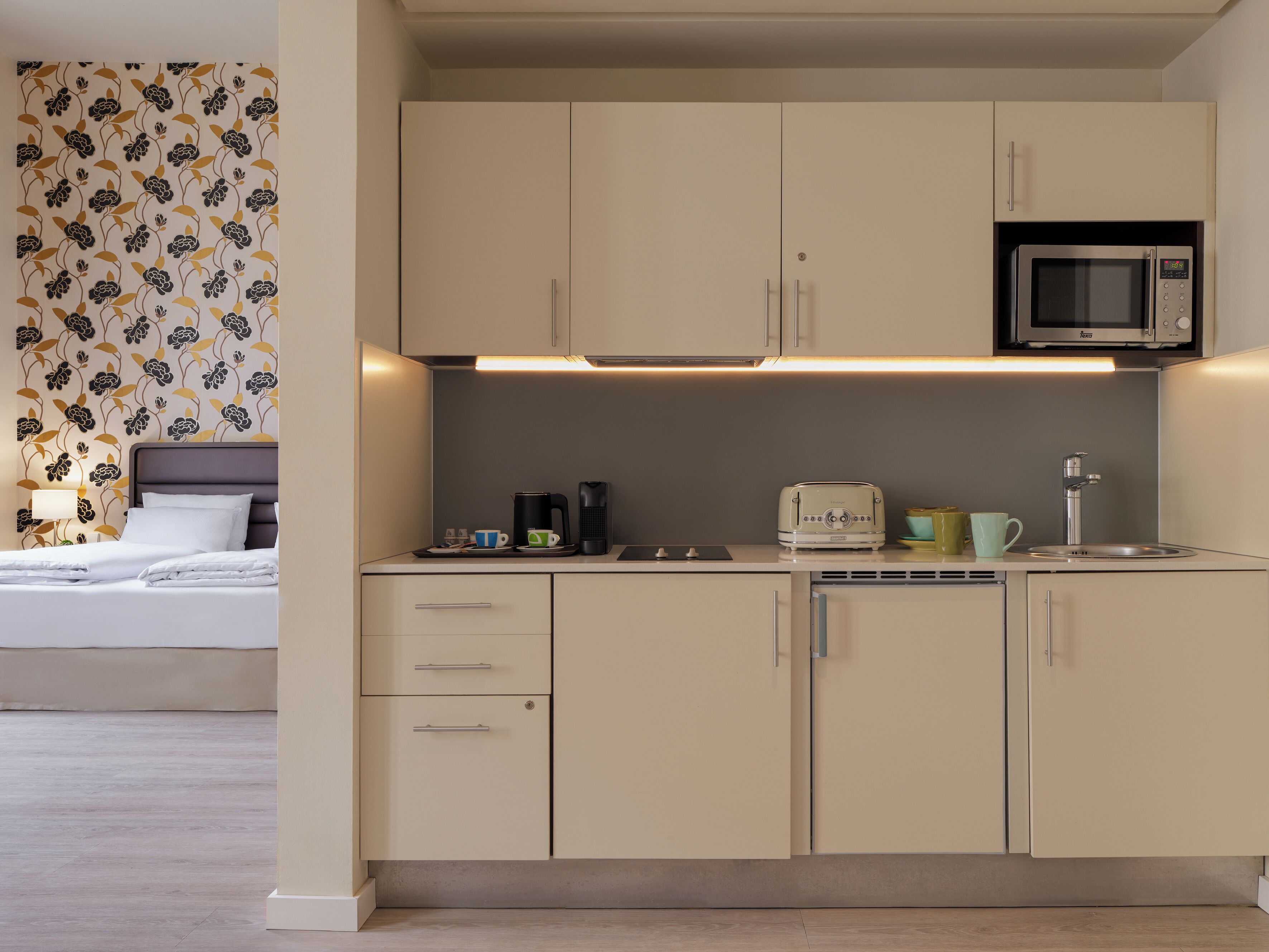 A compact kitchenette with a microwave, toaster, and coffee maker, a bed with a headboard, and a patterned wallpaper.