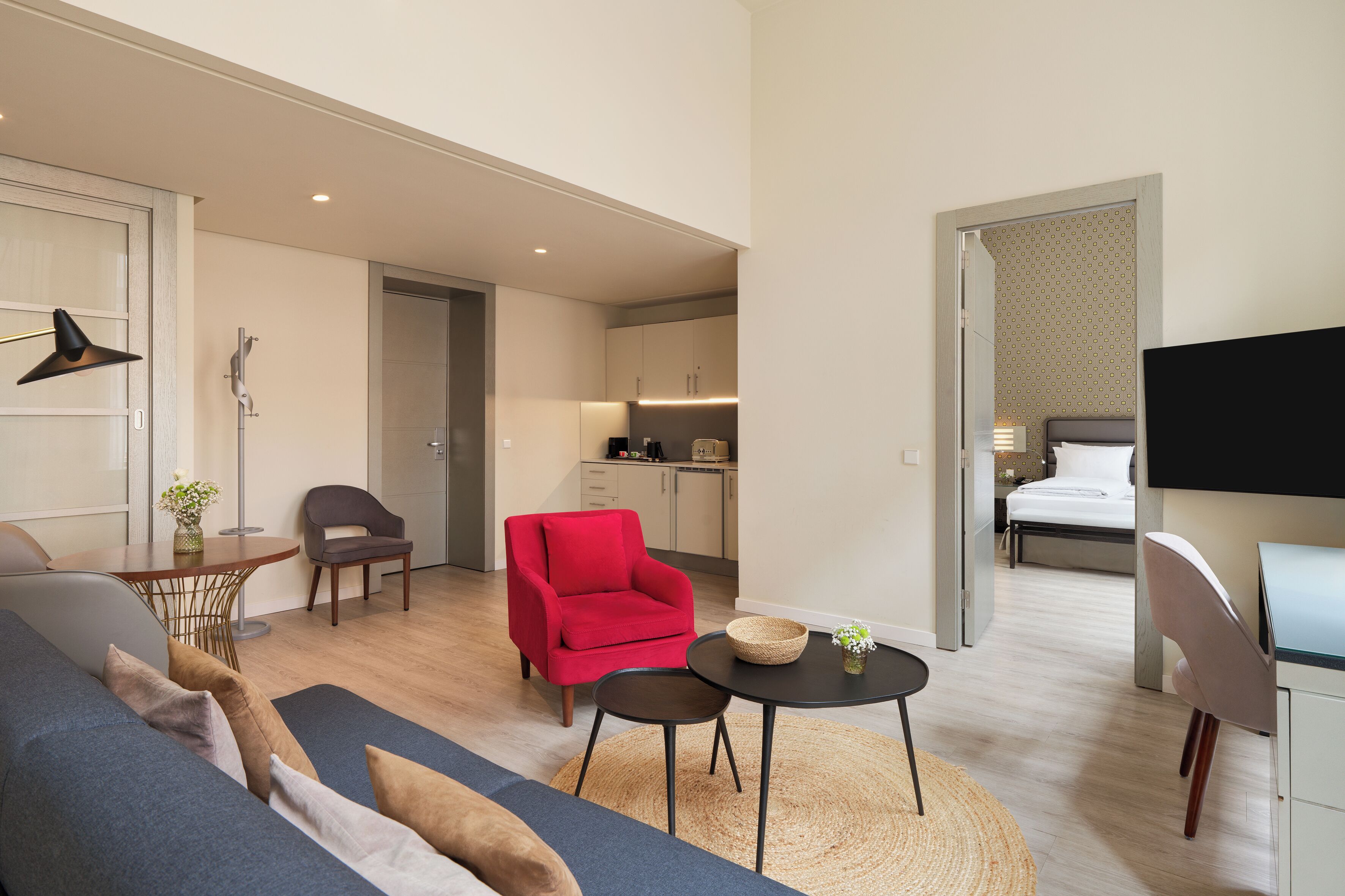 Una habitación de hotel moderna con sofá, un sillón rojo, una mesita, televisor y una cocina al fondo.