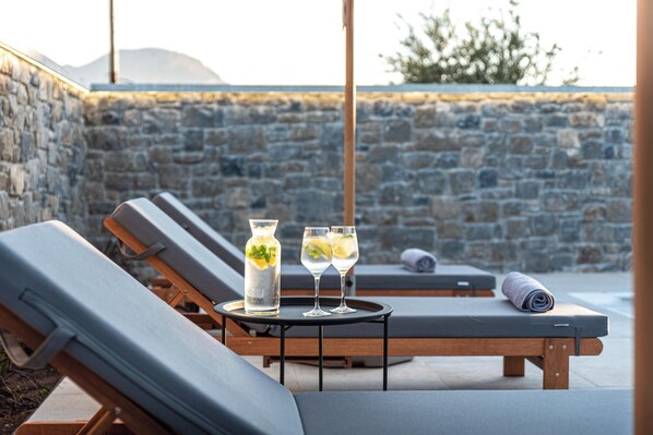 A sunlit outdoor lounge area with stone walls, two lounge chairs, a table with a glass pitcher and two glasses, and a rolled towel.