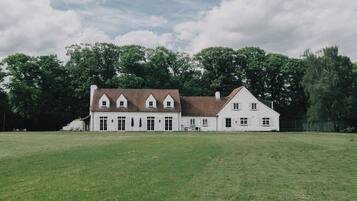 A large, white building with a tiled roof and multiple windows, surrounded by a grassy field and trees.