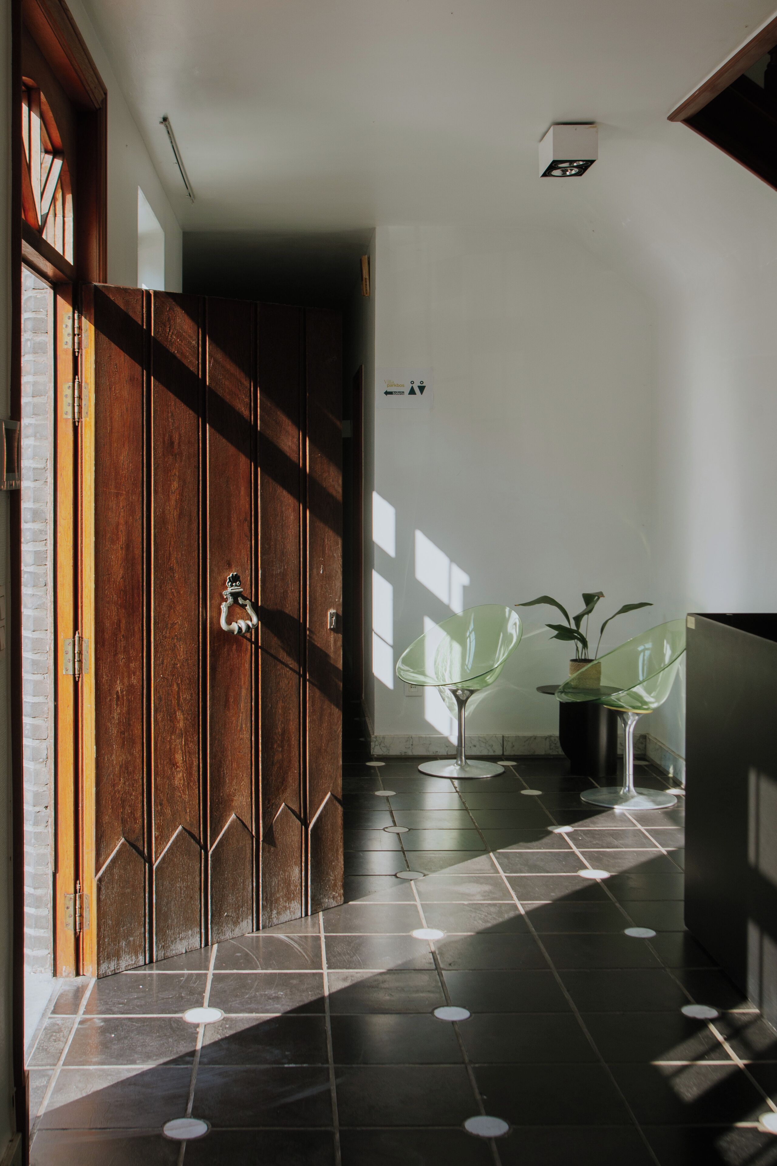 A hallway with a wooden door, a tiled floor with circular patterns, and two glass chairs with plants.