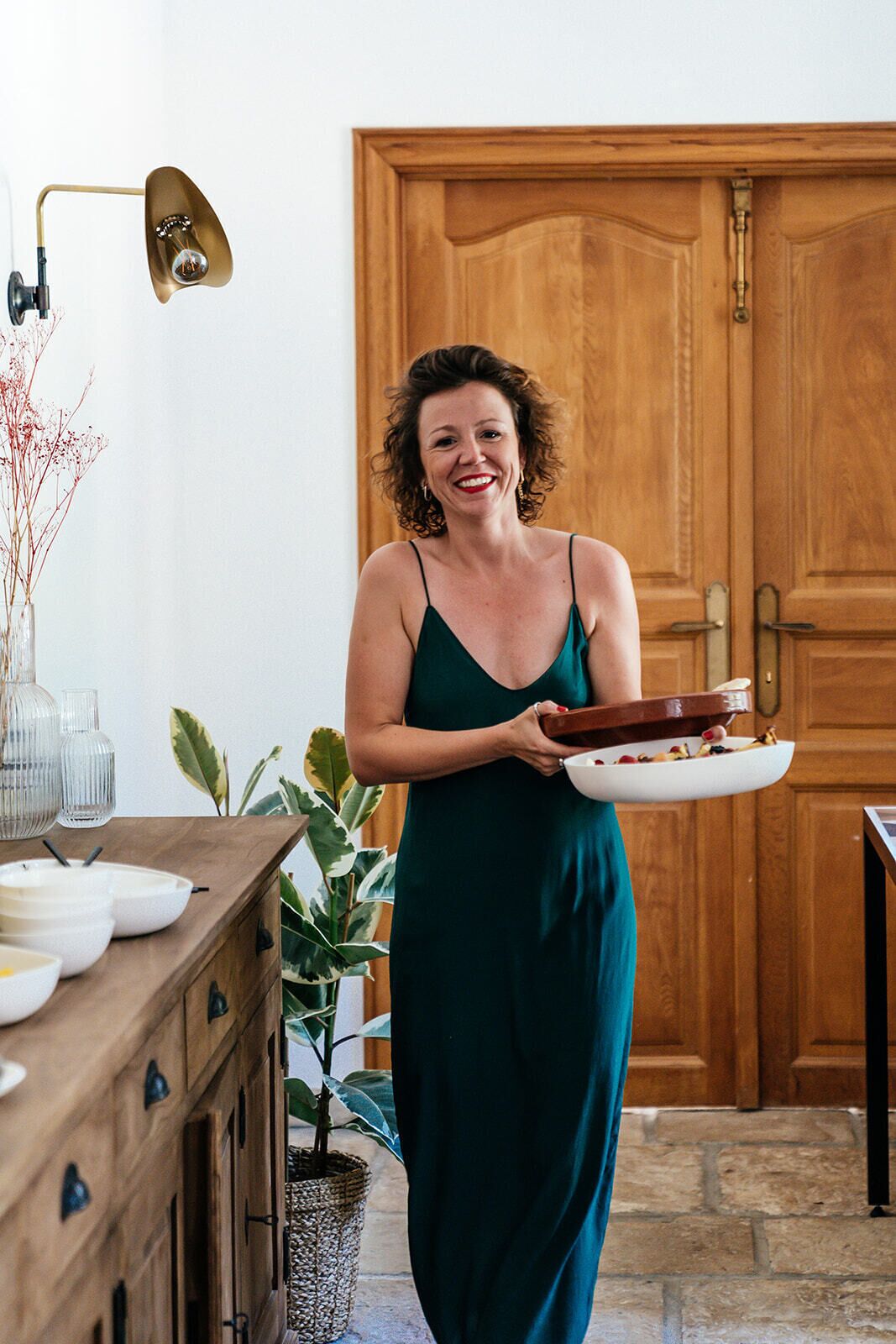 A woman in a green dress holding a white plate with food, standing in a room with wooden cabinets, a plant, and a door in the background.