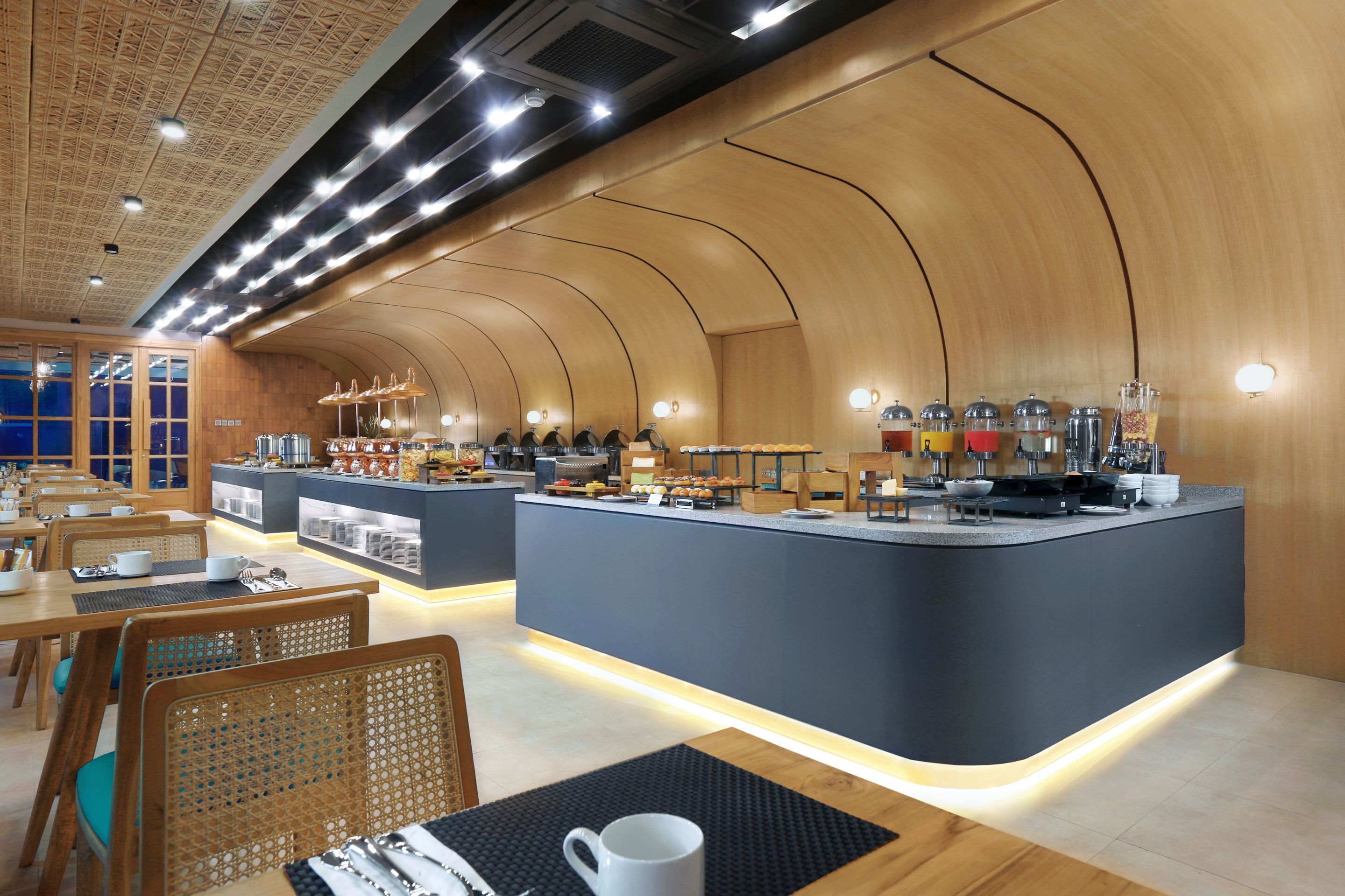 Um café moderno com paredes curvas de madeira, um balcão central com diversos alimentos e áreas de estar com mesas e cadeiras.