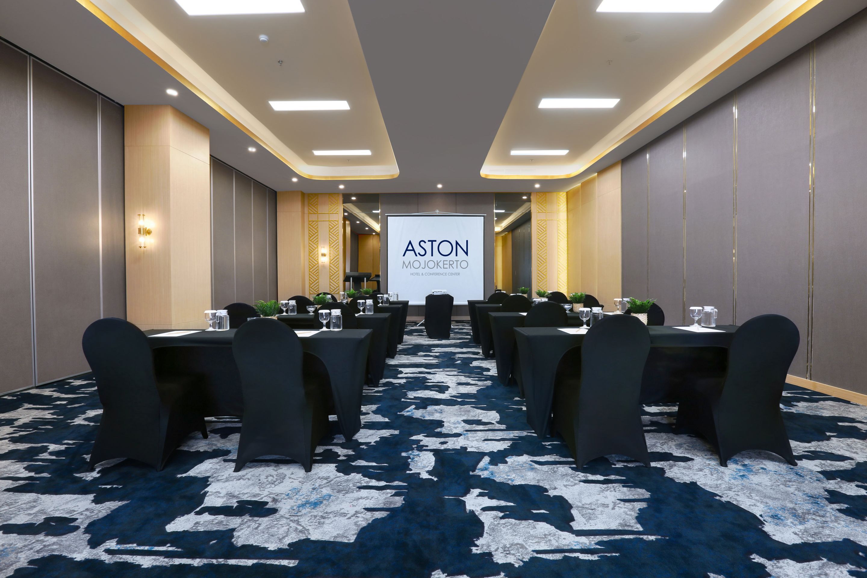 A conference room with a large screen displaying "ASTON MOJOKERTO HOTEL & CONFERENCE CENTER" at the front. Tables are arranged for meetings, with chairs and water glasses on each.