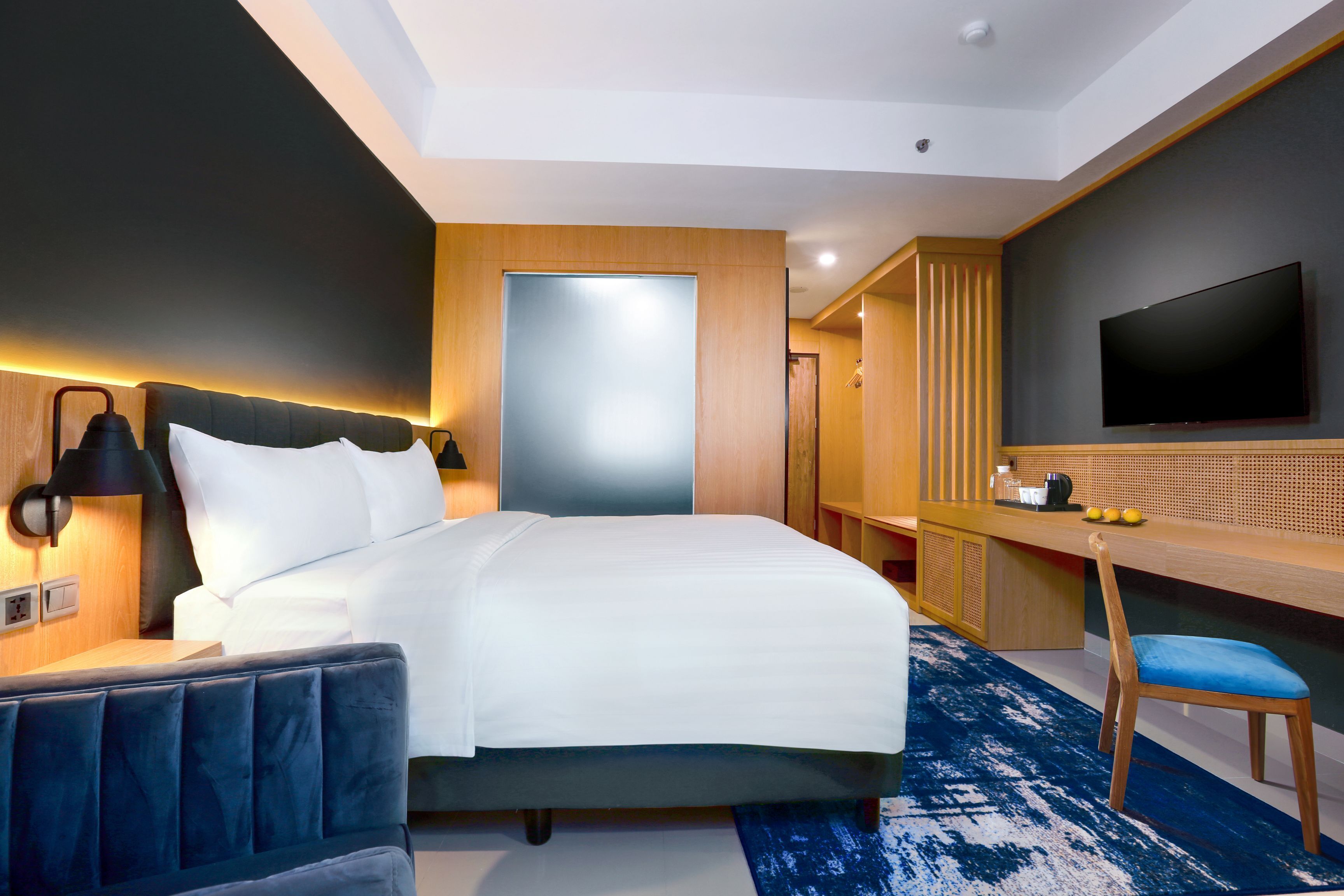 Quarto de hotel com cama, uma escrivaninha com cadeira, televisão na parede e uma janela grande.