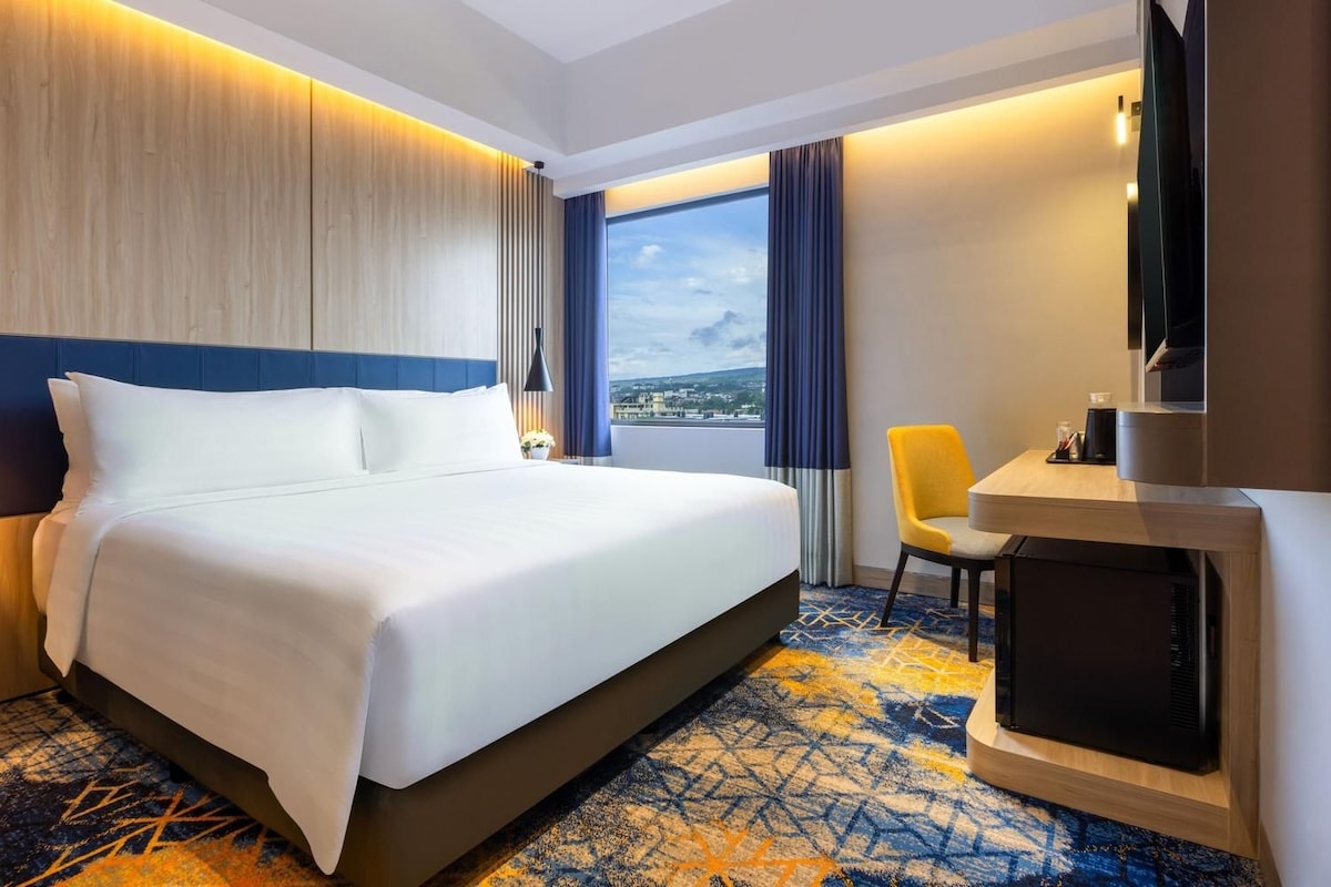 A hotel room with a large bed, a desk with a chair, a television, and a view of the city.