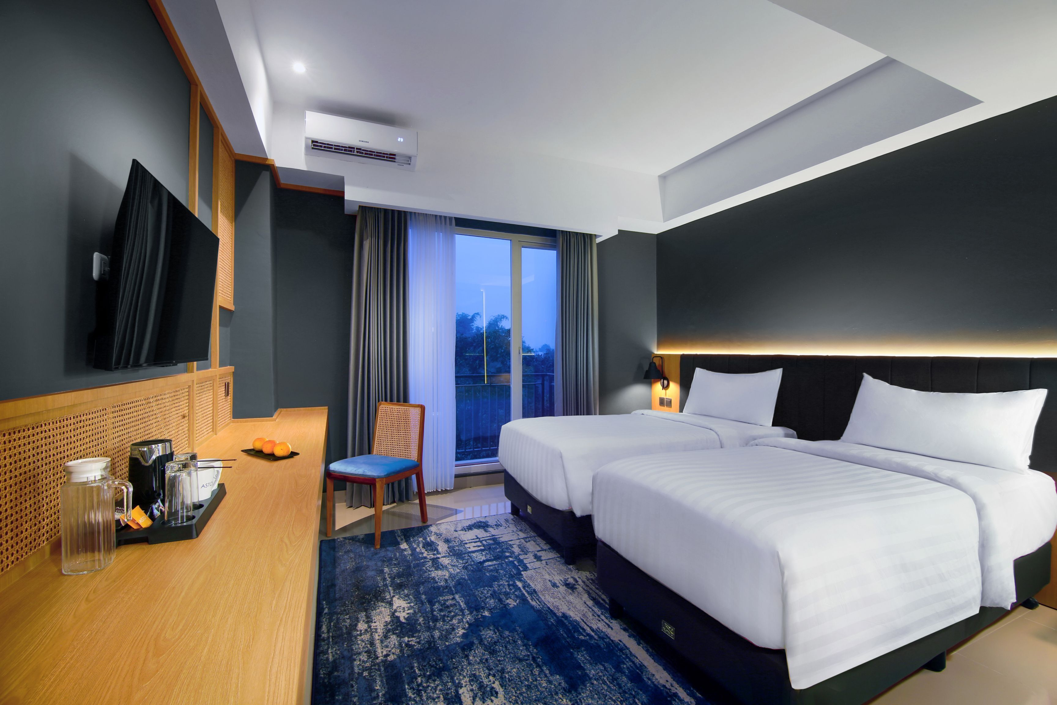 A hotel room with two beds, a TV, a desk with a coffee maker, and a view of the outdoors.