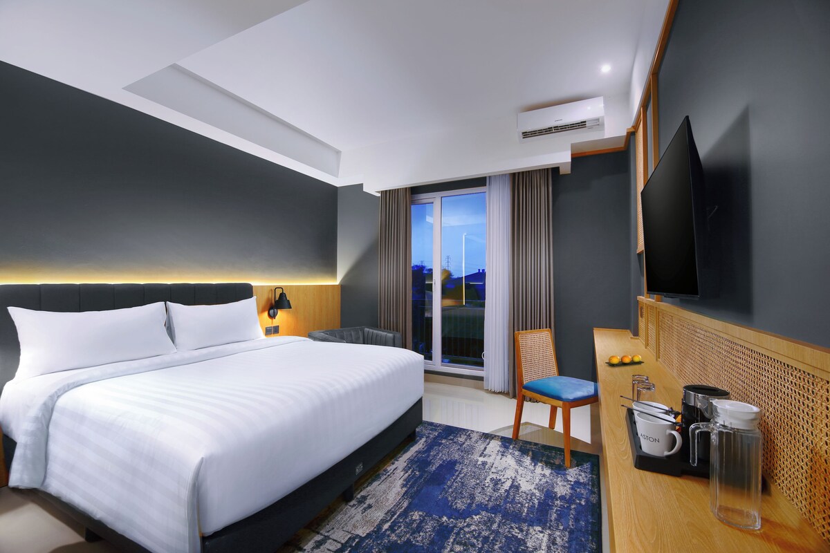 A modern hotel room with a large bed, a desk with a chair, a flat-screen TV, and a view of the outdoors.