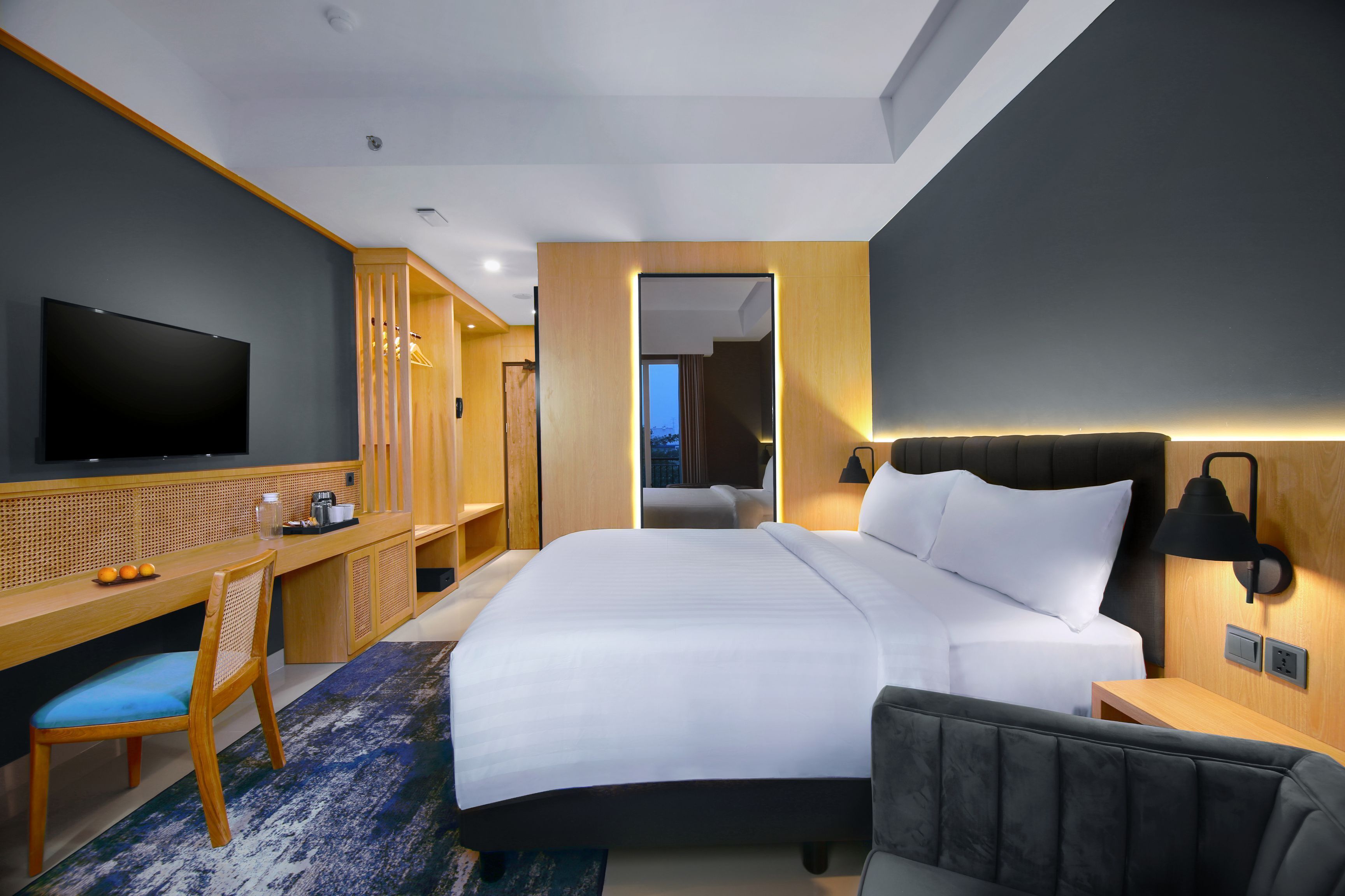A modern hotel room with a large bed, a desk with a flat-screen TV, a chair, and a bedside table with a lamp.