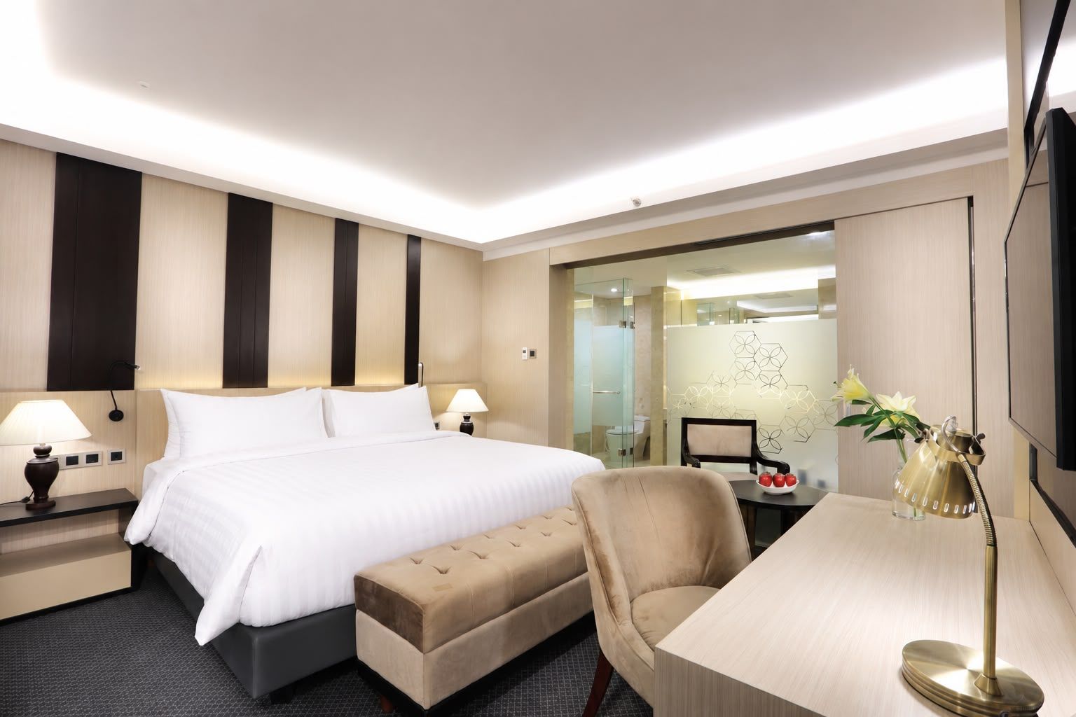 Quarto de hotel com cama grande, mesas de cabeceira com luminárias, escrivaninha com cadeira e divisória de vidro separando o quarto do banheiro.