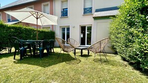 Une terrasse avec une table et des chaises, un grand parasol, et un bâtiment doté de fenêtres et d’une porte.