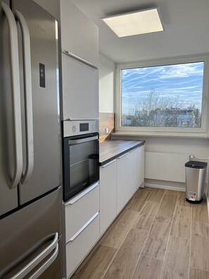 A modern kitchen with a built-in oven, a stainless steel refrigerator, and a window with a view of the sky and trees.