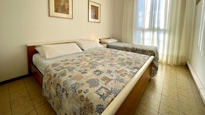 Quarto de hotel com uma cama grande, duas camas menores, uma janela com cortinas e duas fotos emolduradas na parede.