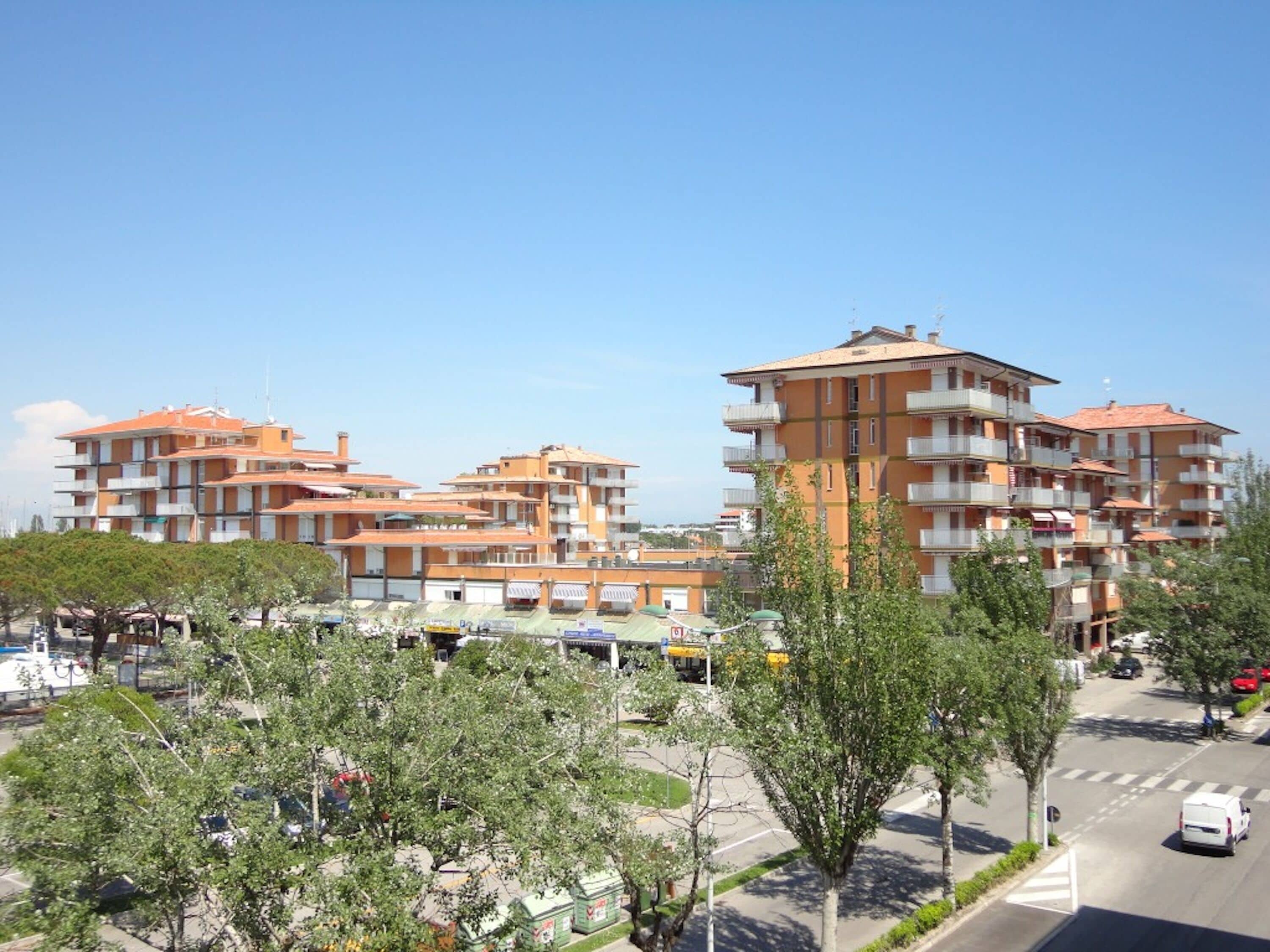 A multi-story residential building with balconies, a parking area, and trees in the foreground.