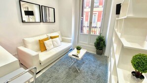 A modern living room with a white sofa, a small coffee table, and a grey rug. There are two framed pictures on the wall, a window with a view of buildings, and a potted plant on the floor. - Design apartment in Plaza Mayor 2 I (Madrid)