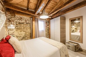 Et soveværelse med træloft, stenvægge, en seng med hvide sengetæpper, en rød pude, en hængende lampe og et spejl på væggen.