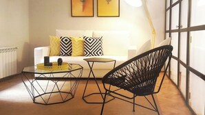 Una sala moderna con un sofá blanco, almohadones estampados en amarillo y negro, una mesa de centro de vidrio, una silla de estructura metálica negra y dos obras de arte con piñas enmarcadas en la pared.